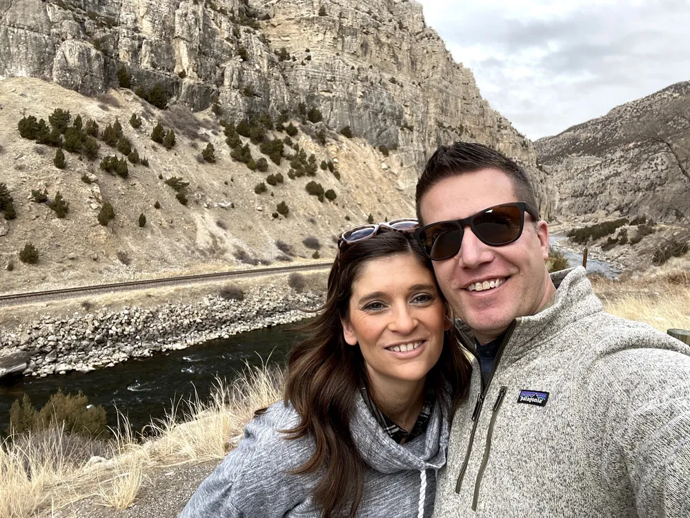 Emily and Josh cheek to cheek by a creek in a canyon, pale limestone cliffs rising behind them