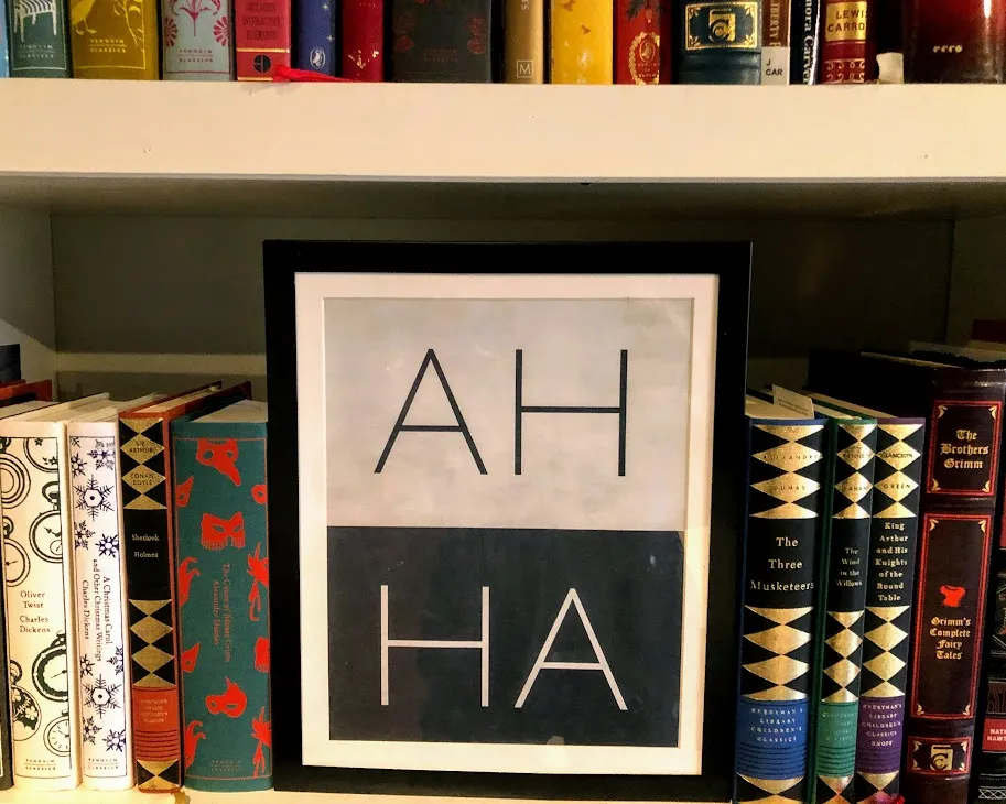 A framed print reading AH HA standing on a bookshelf between rows of hardback classics