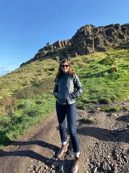 Emily on the dirt path up Arthur's Seat, grassy slope and rocky crags behind her under a clear blue sky