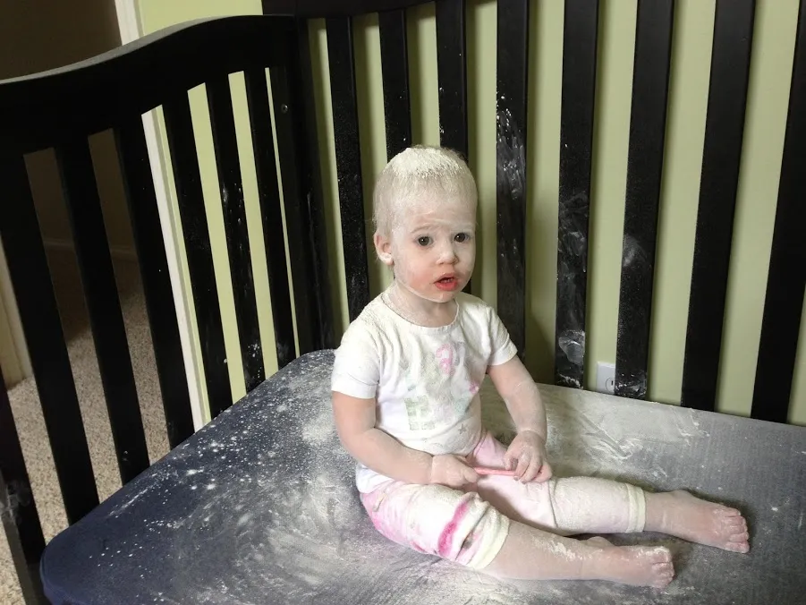 A toddler sitting in her cot covered head to toe in spilled baby powder, looking straight at the camera