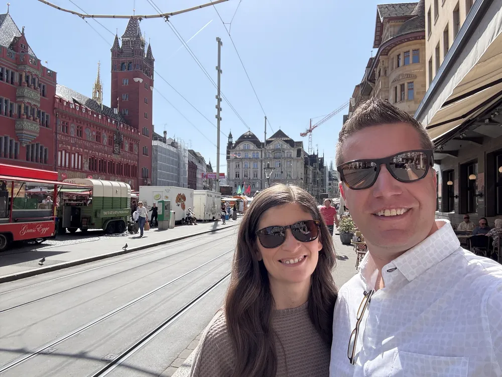 Emily and Josh on a Basel street by the tram tracks, the red sandstone town hall and market stalls behind them
