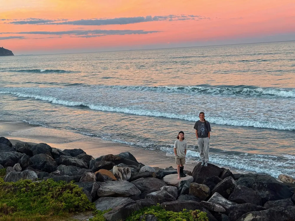 Mia and Case standing on the boulders at the tide line, the sky above the sea burning orange and pink at sunset