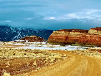 A dirt road curving towards banded red and white bluffs, snow in the hollows and dark mountains behind
