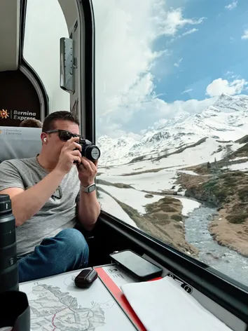 Josh raising a camera to the window of the Bernina Express, a snowy pass and a meltwater stream running past outside