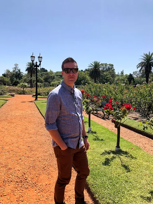 Josh in a garden of red climbing roses, palm trees and a classical lamp post behind him