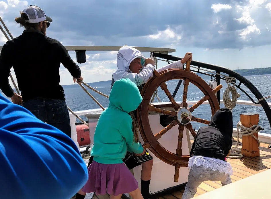 Three of the children taking turns at the wooden ship's wheel on a tall ship, the lake and rigging around them