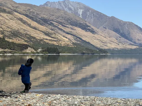 Case at the stony edge of a mirror still lake, mid throw, the golden mountainside reflected across the whole surface
