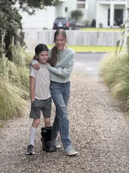 Mia with her arm round Case on a gravel drive between tussock grasses, Case in a backwards cap and a walking boot