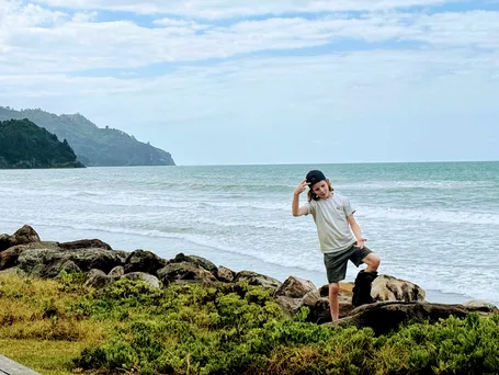 Case posing on the rocks above Waihi Beach in a backwards cap and walking boot, surf breaking behind him and a green headland beyond