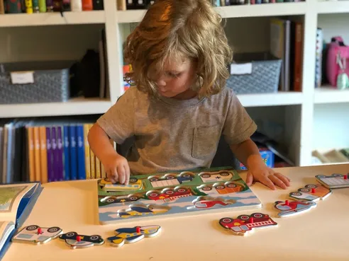 Case at the schoolroom table fitting a wooden vehicle puzzle together, shelves of books behind him