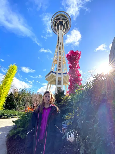 One of the girls in the Chihuly garden, a tall pink glass sculpture and the Space Needle rising behind her