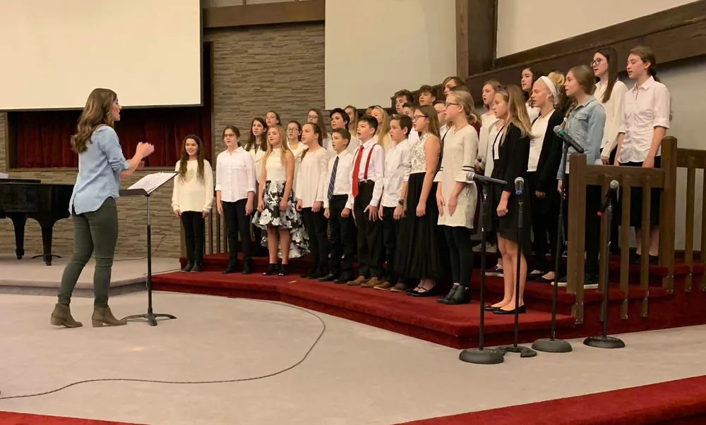 Emily conducting a middle school choir from a music stand, rows of students in black and white singing on the risers