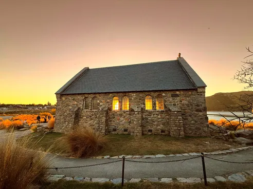 The stone Church of the Good Shepherd at Lake Tekapo at sunset, its windows lit gold from inside and tussock grass all around