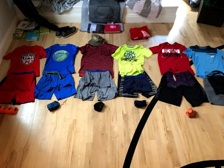 Six outfits laid out in rows on a wooden floor, each a shirt with shorts beneath it, shoes and a cap alongside