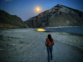 Emily walking a gravel shore towards the Buffalo Bill reservoir at dusk, a full moon over the mountain and its light on the water
