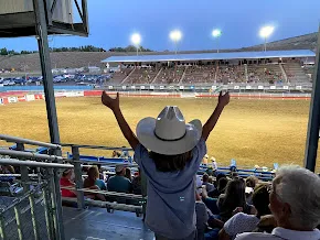 Case in a white cowboy hat with both arms raised, looking down over the floodlit rodeo arena