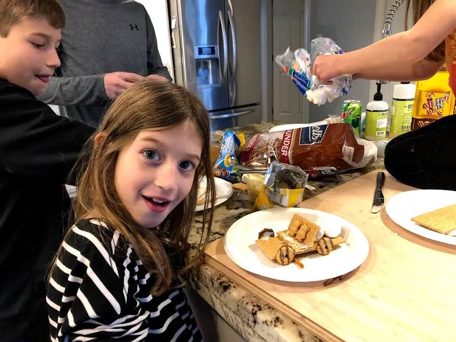 One of the girls grinning beside a paper plate holding a covered wagon built from graham crackers and marshmallows