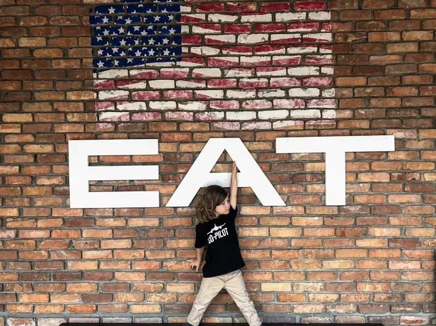 The youngest reaching up to touch the big white letters spelling EAT on a brick wall, a painted flag above