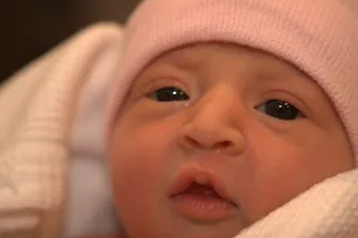 Evie as a newborn in a pink hospital hat, dark eyes wide open looking at the camera