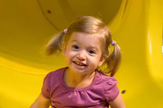 Evie as a toddler in bunches and a purple top, grinning at the bottom of a yellow tube slide