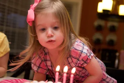 Evie in a pink bow and checked dress leaning in to blow out three birthday candles