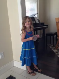 Evie in a blue sundress and sandals holding a small purse, the piano behind her