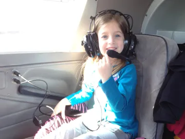 Evie in an oversized headset in the back of a small aeroplane, holding the microphone to her mouth