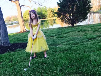 Evie in a yellow princess dress and a golf glove, lining up a putt on the lawn by the pond