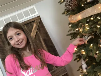 Evie in a pink jumper hanging a glittery tiara ornament on the lit Christmas tree
