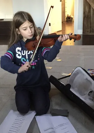 Evie kneeling on the carpet playing the violin, practice sheets spread on the floor around her