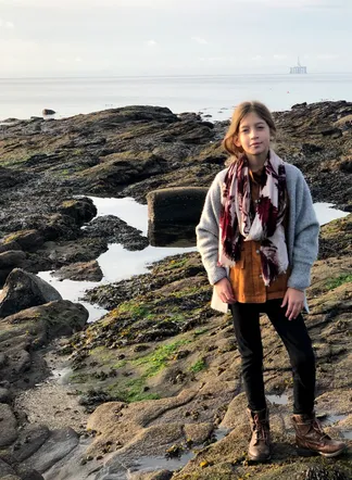 Evie in a scarf and grey cardigan standing on the rocky shore at low tide, a rig out on the flat sea behind her