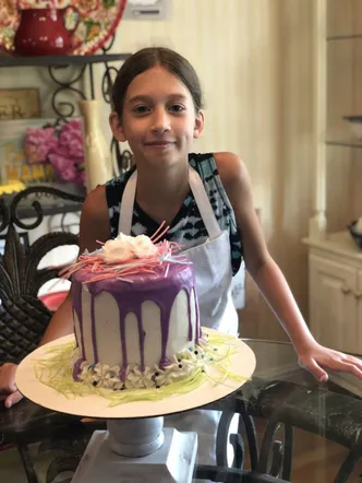 Evie in an apron holding up a layer cake she has made, purple icing dripping down the white frosting