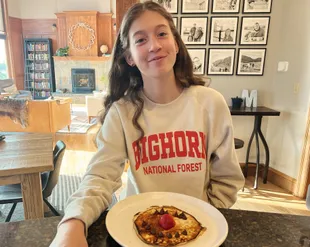 Evie in a Bighorn National Forest sweatshirt holding out a pancake decorated with chocolate chips and a strawberry