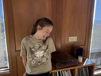 Evie laughing with her tongue out in a longhorn and daisies t-shirt, the record player on the table beside her