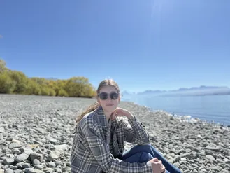 Evie in sunglasses and a checked shirt sitting on a stony lake shore, mountains hazy across the water