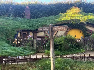 A wooden signpost reading East Farthing and West Farthing in front of a hobbit hole with a yellow round door and a turf roof
