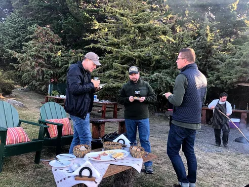 Three men talking while standing around a food table with appetizers, green forest behind them