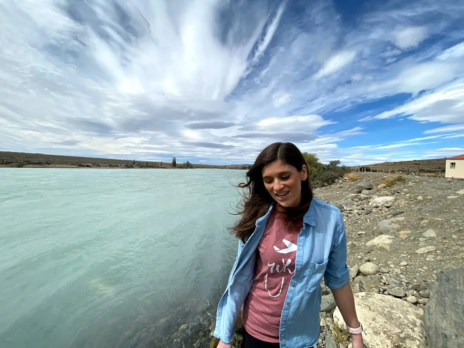 Emily laughing, head turned down, beside a pale turquoise glacial river under a big streaked sky