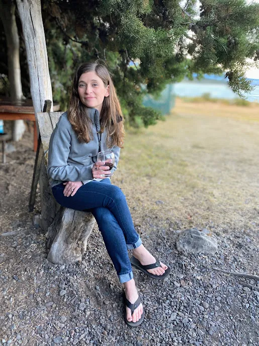 Emily sitting on a tree stump under low branches in Patagonia, a glass in her hand and the lake behind her