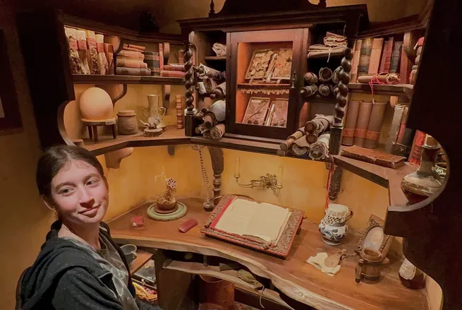 Evie turning to the camera in Bilbo's study, the desk in front of her with an open illustrated book and the shelves behind packed with books and scrolls