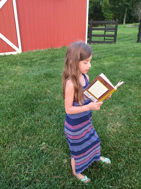 Evie walking across the grass in a striped maxi dress, reading a book, the red barn behind her