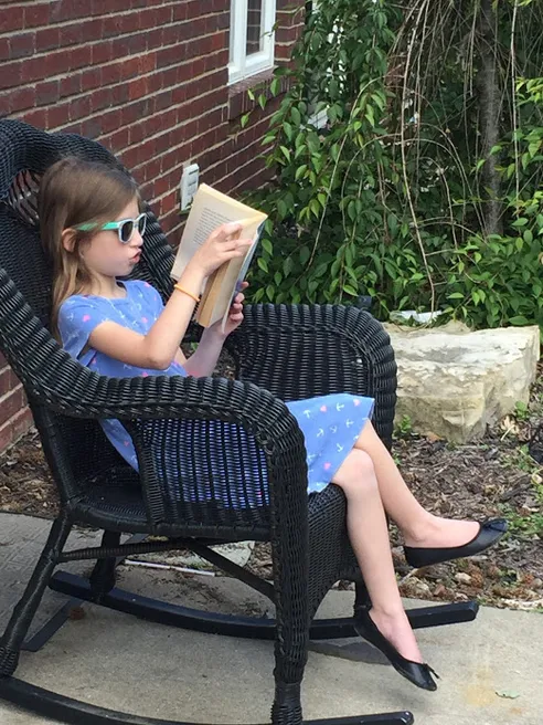Evie in sunglasses curled into a black wicker rocking chair outside, absorbed in a book