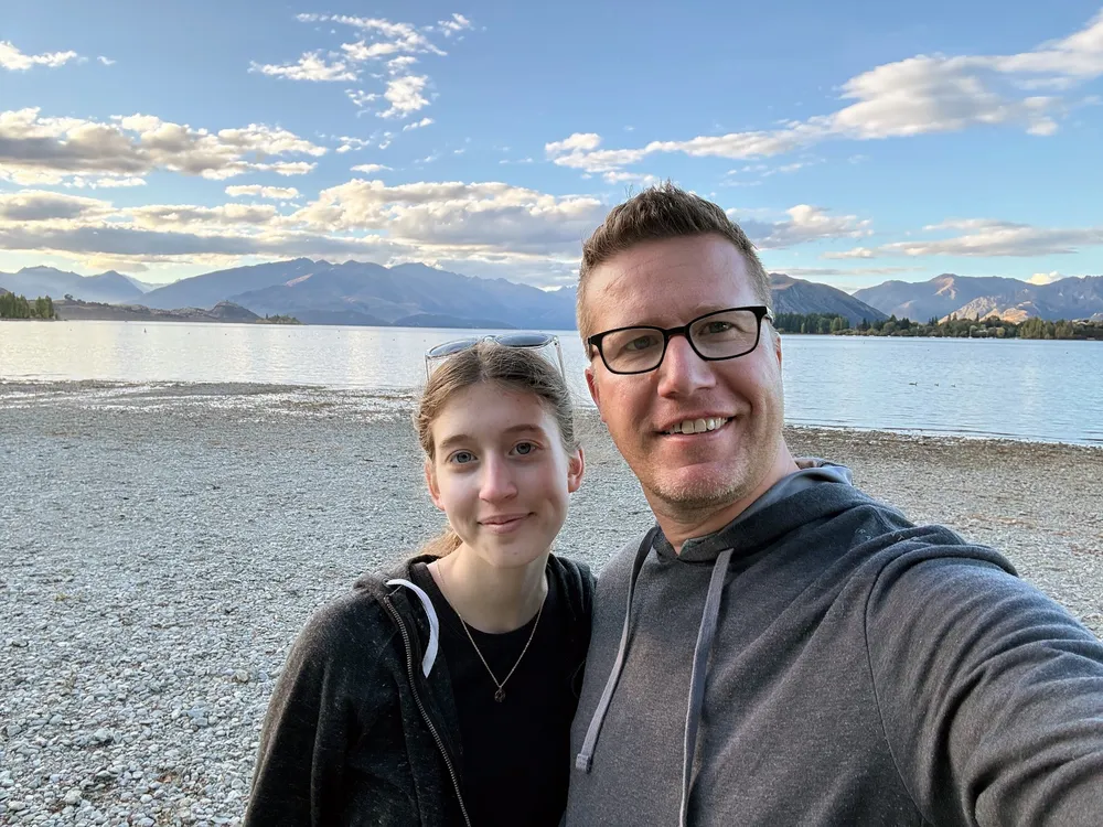 Evie and Josh cheek to cheek on the pebble shore of Lake Wanaka at golden hour, the mountains low behind them