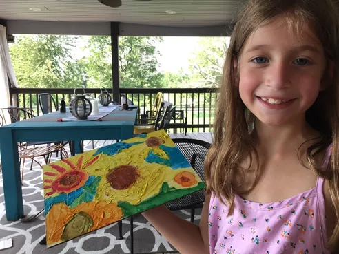 Evie holding up her thickly painted sunflower canvas on the back porch