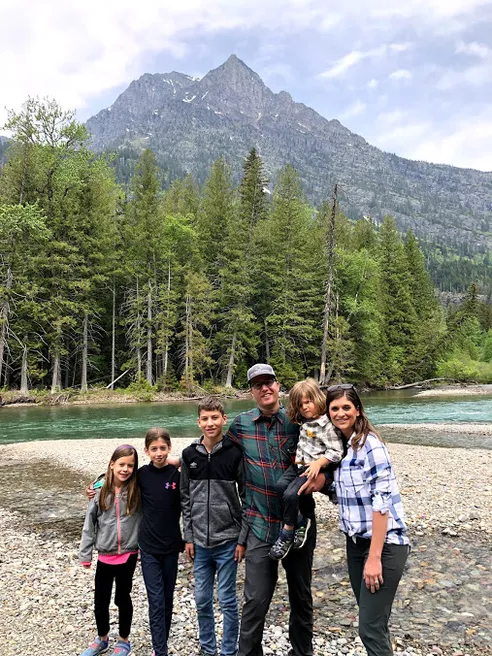 The six of them on the gravel bank of a green river in Glacier National Park, a sharp peak behind the firs