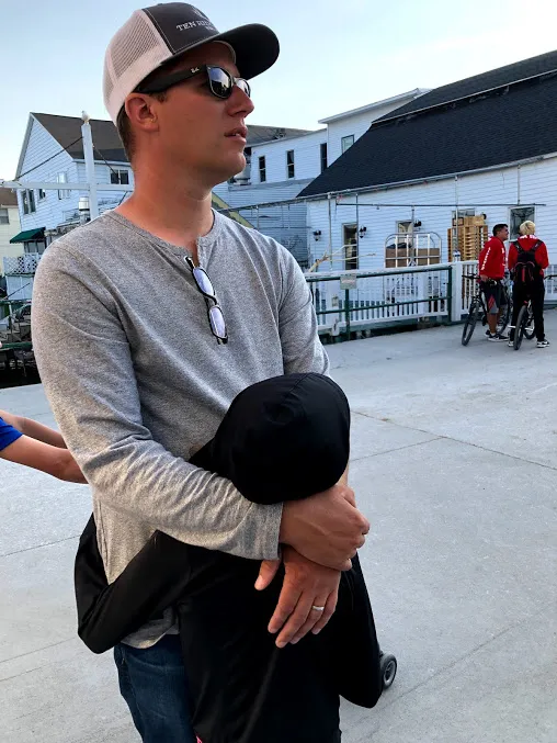 Josh in a cap and sunglasses holding his daughter close against his chest on the street, bikes and buildings behind