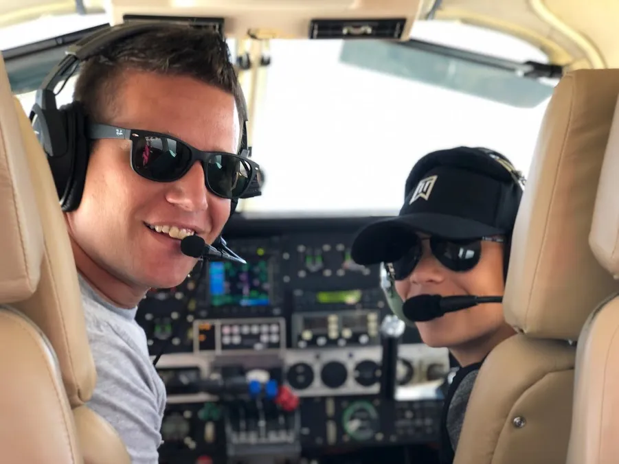 Josh and Preston in the cockpit in headsets and sunglasses, both turned round to the camera