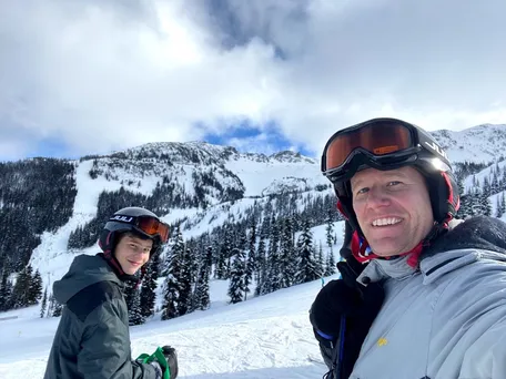 Josh and Preston in helmets and goggles on the piste, snow covered peaks and firs behind them