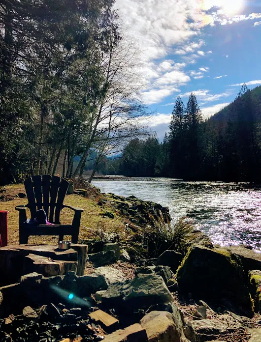 An Adirondack chair and a stone fire pit on the bank of a river, sunlight scattering off the water