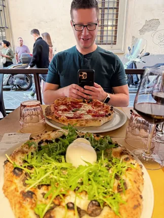 Two Neapolitan pizzas on a pavement table, one heaped with rocket and mozzarella, Josh photographing his own across the table